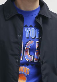 Giacca nera con bottoni sopra una maglietta blu con testo audace e una grafica di un pallone da basket con motivi colorati.