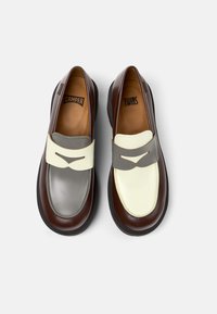 Mocasines bicolor con cuero en marrón, gris y crema. Presentan una amplia correa con un detalle ranurado. Punta redonda y suela gruesa negra.