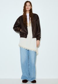 Braune Leder-Bomberjacke, cremefarbenes asymmetrisches Top mit Spitzenbesatz, hellblaue Jeans mit weitem Bein und dunkle Fell-Slipper.