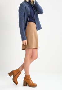 Cárdigan azul de punto, falda beige y botines marrones de tacón. El cárdigan tiene un frente abierto; los botines cuentan con un tacón grueso y cremallera.
