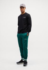 Felpa nera con logo Calvin Klein. Pantaloni della tuta verdi con strisce laterali e logo. Scarpe da ginnastica nere e grigie. Cappellino grigio.