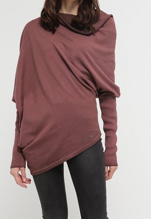 Mujer con un suéter asimétrico color malva con mangas largas ajustadas y pantalones oscuros sobre un fondo liso.