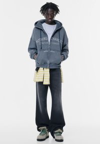 Kapuzenpullover in verblasstem Marineblau mit dem Text "NICHTS ÄNDERT SICH", über einem gelb gestreiften Shirt getragen, kombiniert mit weit geschnittenen dunklen Jeans und grauen Schuhen.