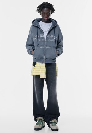 Sweat-shirt à capuche en bleu marine délavé avec le texte "RIEN NE CHANGE", superposé à une chemise à rayures jaunes, associé à un jean large sombre et des chaussures grises.