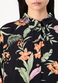 Dunkelbluse mit floralem Muster, das lebendige orange Blumen und grüne Blätter auf einem schwarzen Hintergrund zeigt. Abgeschlossen mit einer Knopfleiste und Kragen.