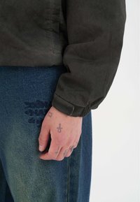 Mørkegrøn, tekstureret jakkeærme med elastisk kant ved siden af lyseblå denim, der har præget tryk. Synlig hånd med tatoveringer hviler på buksebenet.