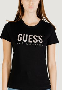 T-shirt in cotone nero con una grafica in glitter argento "GUESS LOS ANGELES" sul petto, scollatura rotonda e maniche corte.