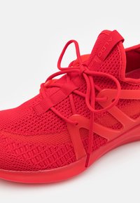 Röd atletisk sko gjord av texturerad stickad tyg med ett flexibelt plastöverdrag. Har runda snören och en vadderad sula.