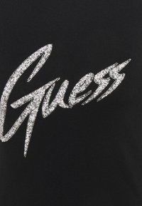 Logo "Guess" napsané kurzívou se stříbrnými kamínky na černém textilním pozadí.