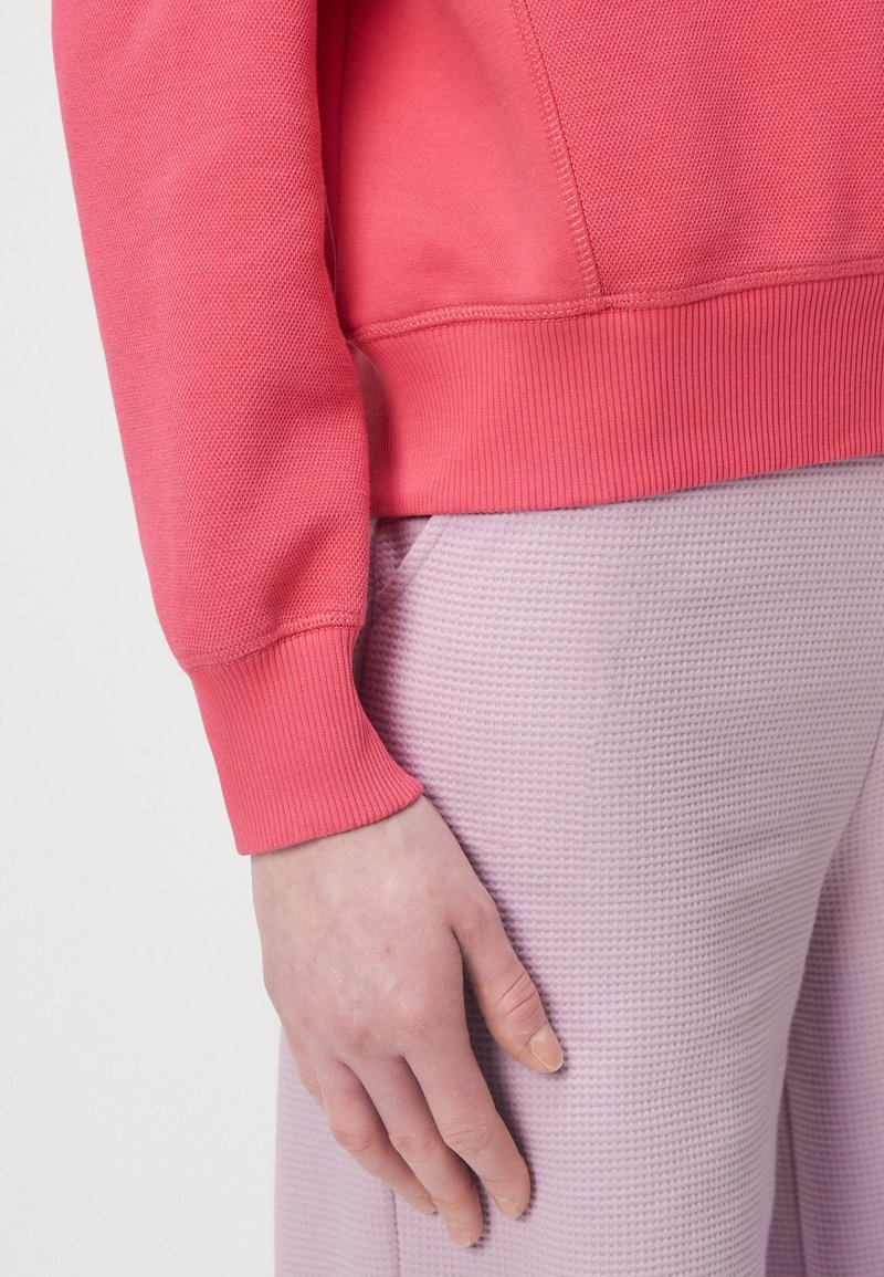 Inquadratura ravvicinata di una mano appoggiata su pantaloni texturizzati di un viola chiaro e una felpa rosa a maniche lunghe con polsini e orlo a coste.