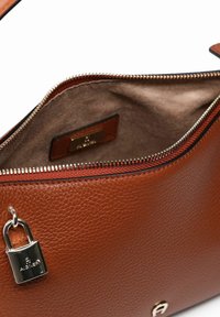 Bruine leren handtas met een ritssluiting, voorzien van een zachte binnenvoering, een metalen hangslot als accent en een subtiele textuur overal.