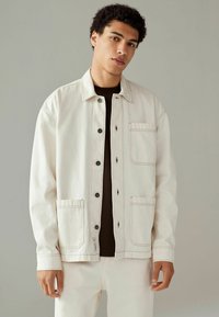 Chaqueta beige claro con cierre de botón, dos bolsillos frontales y costuras visibles. Confeccionada con un tejido duradero y un corte relajado.