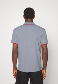 Polo gris avec col, comportant des accents rouges sur le col et les manches. Le tissu semble lisse et léger. Aucun motif visible.