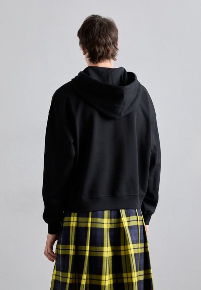 Schwarze, verkürzte Kapuzenjacke mit einer großen Kapuze, kombiniert mit einem karierten Kilt in Gelb und Marineblau. Hat ein einfaches, unauffälliges Design.