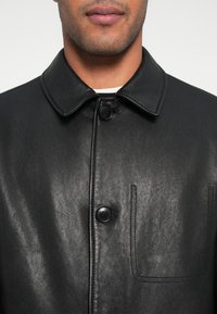 Schwarzer Ledermantel mit klassischem Kragen, zwei Knöpfen und einer aufgesetzten Tasche, glatter Textur mit einem dezenten Glanz.