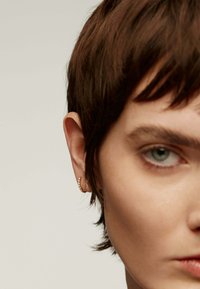 Pendiente de aro dorado con pequeños cristales transparentes, con un diseño curvado, situado sobre un fondo neutro difuminado. Se puede ver cabello corto y oscuro.