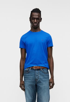 Bărbat cu păr împletit purtând un tricou albastru deschis și blugi albaștri cu o centură maro, stând în fața unui fundal simplu și deschis la culoare.