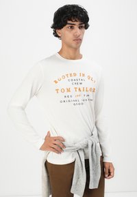 Νεαρός άνδρας με λευκό μακρυμάνικο πουκάμισο με πορτοκαλί κείμενο και γκρι πουλόβερ δεμένο γύρω από την μέση του, μπροστά σε απλό φόντο.