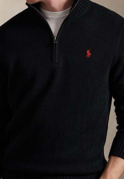 Mann trägt einen schwarzen Strickpullover mit halbem Reißverschluss und einem roten gestickten Logo auf der Brust sowie ein weißes Hemd darunter.