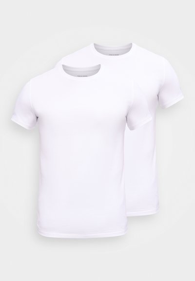 Δύο μονόχρωμες λευκές κοντομάνικες μπλούζες με στρογγυλή λαιμόκοψη, ελαφρώς επικαλυπτόμενες σε ουδέτερο φόντο.