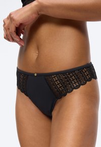 Η μέση γυναίκας με μαύρα εσώρουχα διακοσμημένα με δαντέλα και ένα μικρό διακοσμητικό γούρι στο κέντρο μπροστά. Τα χέρια τοποθετημένα κοντά στη μέση.