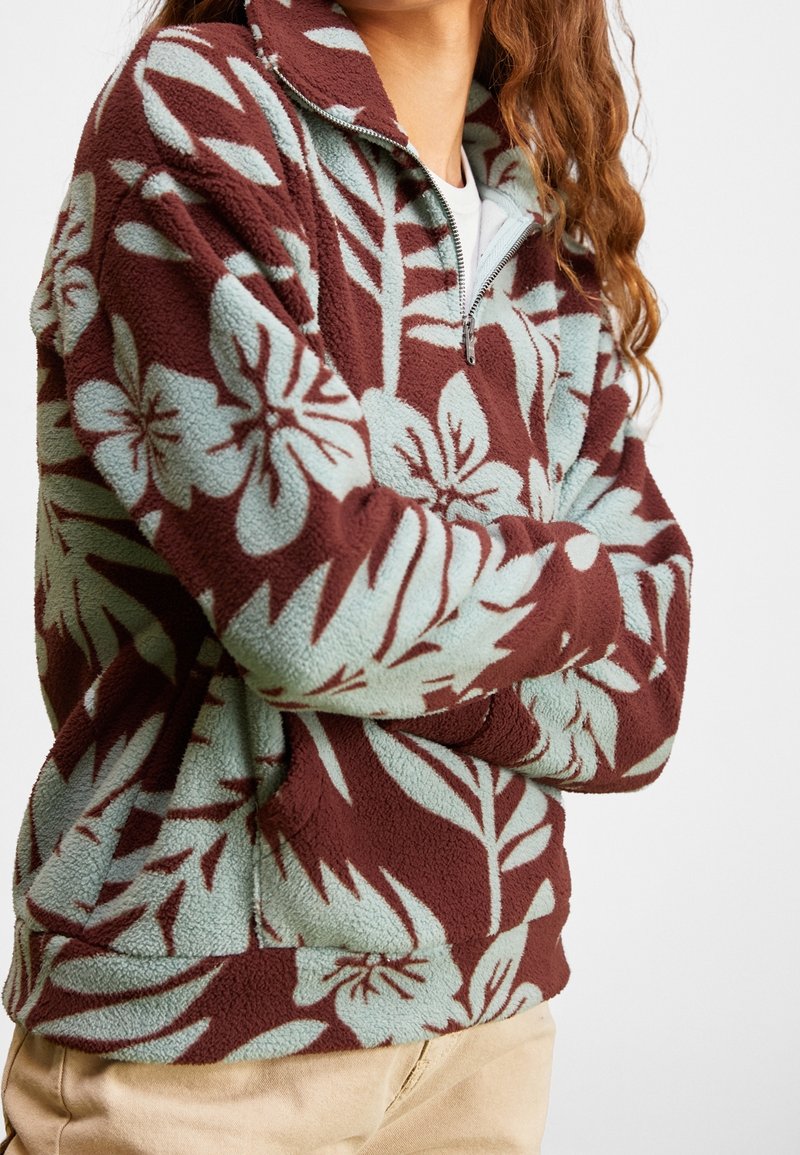 Personne portant un pull polaire marron et bleu clair à motif floral avec un col à demi-fermeture éclair et un pantalon beige, bras croisés.