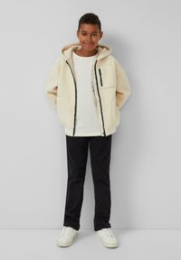 Fleece-futternde, cremefarbene Zip-Jacke mit Kapuze, Vorderentaschen und schwarzen Akzenten, kombiniert mit einem leichten Shirt, dunklen Hosen und weißen Sneakers.