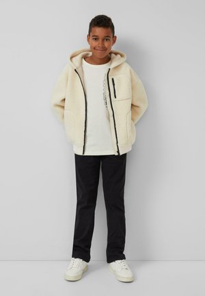 Fleece-futternde, cremefarbene Zip-Jacke mit Kapuze, Vorderentaschen und schwarzen Akzenten, kombiniert mit einem leichten Shirt, dunklen Hosen und weißen Sneakers.