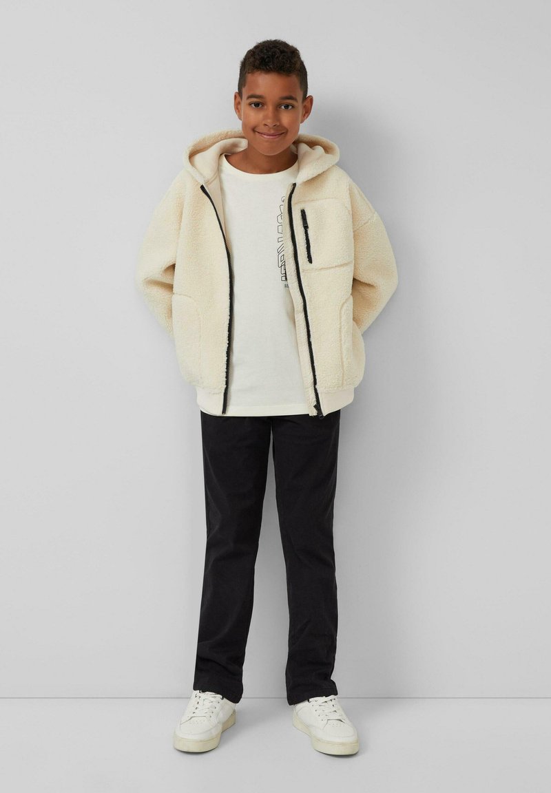 Fleece-futternde, cremefarbene Zip-Jacke mit Kapuze, Vorderentaschen und schwarzen Akzenten, kombiniert mit einem leichten Shirt, dunklen Hosen und weißen Sneakers.