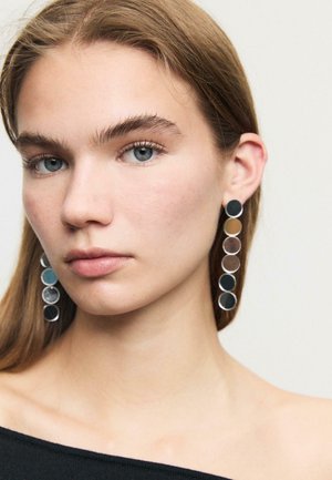 Mladá žena se rovnými světle hnědými vlasy, oblečená v černém topu bez ramínek a dlouhých visících náušnicích s pěti mnohobarevnými cirkulárními diskem.