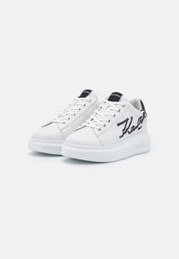 Hvite sneakers med en glatt læroverdel, rund tå og tykk hvit såle. Har svarte logoakkenter og kontrasterende sømdetaljer.