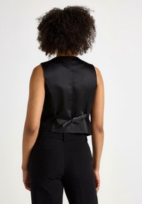 Czarny satynowy kamizelka z wysokim dekoltem, dopasowaną formą i akcentem w postaci kokardy z ty�łu. Noszona na dopasowanych czarnych spodniach, podkreślająca smukłą sylwetkę.