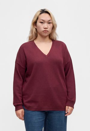 Giovane con lunghi capelli biondi che indossa un maglione largo color granata con scollo a V e jeans blu, in piedi davanti a uno sfondo bianco neutro.