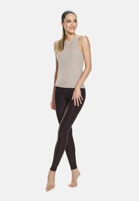 Giovane donna sorridente, in piedi a piedi nudi con le gambe incrociate, indossando un top beige senza maniche e leggings neri su uno sfondo semplice.