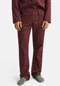 Man wearing red paisley-patterned pajama pants and matching top with gray woolen socks, standing against a white background.