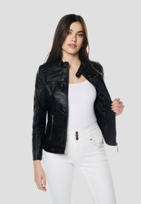 Čierna kožená bunda s stojacím golierom, vreckami na zips a vyšívanými detailmi na rukávoch, nosená otvorená cez biely top a džínsy.