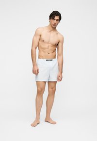 Μυώδης άνδρας όρθιος με γυμνά πόδια, που φοράει λευκά μποξεράκια Moschino με λεπτές κάθετες ρίγες σε απλό ανοιχτό φόντο.