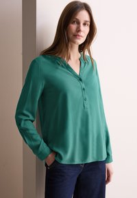 Blusa verde a maniche lunghe con scollatura a V e chiusura con bottoni. Tessuto morbido e liscio con una vestibilità ampia, abbinata a jeans scuri.