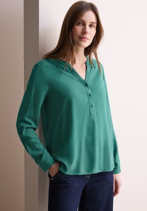 Blouse verte à manches longues avec un décolleté en V et une patte de boutonnage. Tissu doux et lisse avec une coupe ample, associée à un jean foncé.