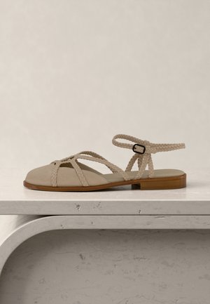 Beige flad sandal med flettede stropper, ankelspænde og træsål, placeret på en lys stenovertflade mod en neutral baggrund.