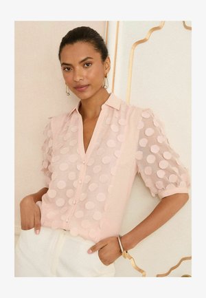 Hele roosa pluus, millel on nööbid ees ja tekstuurilised, ringikujulised puffdetailid varrukatel. Sile kangas ja mugav lõige.