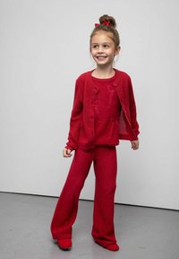 Czerwony dziergany kardigan na guziki, nałożony na pasujący sweter i rozszerzane spodnie. Miękka tekstura, spójna kolorystyka i minimalistyczne detale.