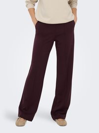 Personne portant un pantalon large violet foncé, un sweat-shirt beige et des ballerines beiges, les mains dans les poches, sur un fond clair.