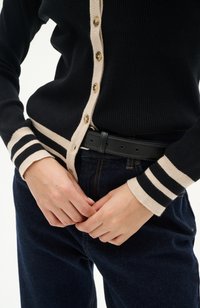 Črn rebrast cardigan z bež gumbi, črtastimi manšeti, obdan z temno modrimi jeans hlačami in črnim usnjenim pasom.