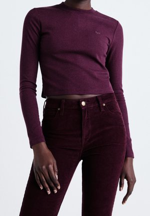 Personne à la peau foncée portant un crop top violet à manches longues côtelé et ajusté, ainsi qu'un pantalon en velours côtelé violet taille haute, debout avec une main détendue.
