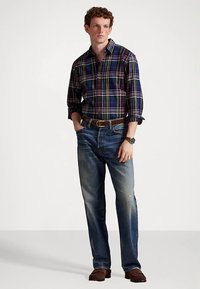 Koszula w kratę w kolorze granatowym z zielonymi, żółtymi i czerwonymi akcentami, zestawiona z jasnoniebieskimi dżinsami. Brązowy pasek i zegarek dopełniają strój.