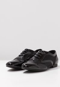 Chaussures noires à lacets avec un bout en cuir verni brillant et des côtés en tissu texturé, présentant des détails de brogue et un talon bas.