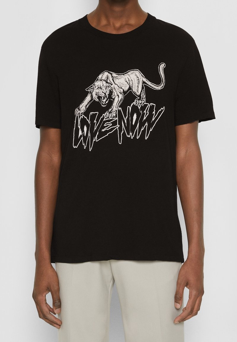 Czarny t-shirt z białą ilustracją warczącej pantery nad wyrazistym, poszarpanym napisem "LOVE NOW", noszony przez osobę w beżowych spodniach.