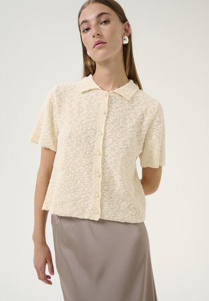 Cream-colored short-sleeve knit cardigan with a collar, featuring a textured fabric and pearl buttons down the front. Worn over a smooth skirt.