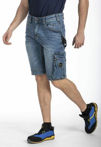 Shorts cargo en denim bleu clair avec plusieurs poches, des détails cousus et un patch logo. Portés avec des chaussures de sport noires et bleues.